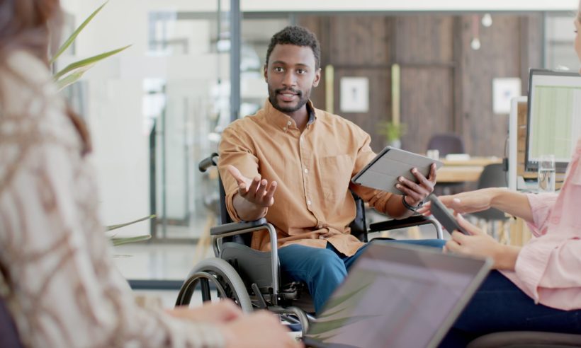 Professional in a wheelchair speaking with colleagues in a modern office meeting environment.