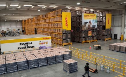 A modern DHL warehouse. In the foreground, a worker walks across the floor pulling a pallet jack near stacks of shrink-wrapped boxes. Tall orange shelving units filled with goods line the background, with yellow DHL banners and a prominent “ESCUELA DE EXCELENCIA” wall on the left. The space is clean, brightly lit by skylights, and appears highly organized.
