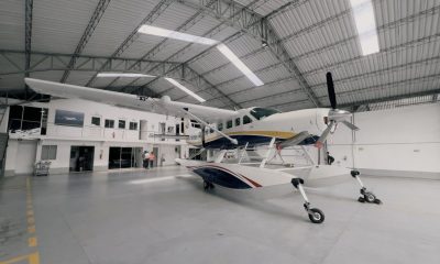 A large white amphibious seaplane (Cessna Caravan-type aircraft with floats) is parked inside a spacious modern hangar. (Photo: Alma Air)