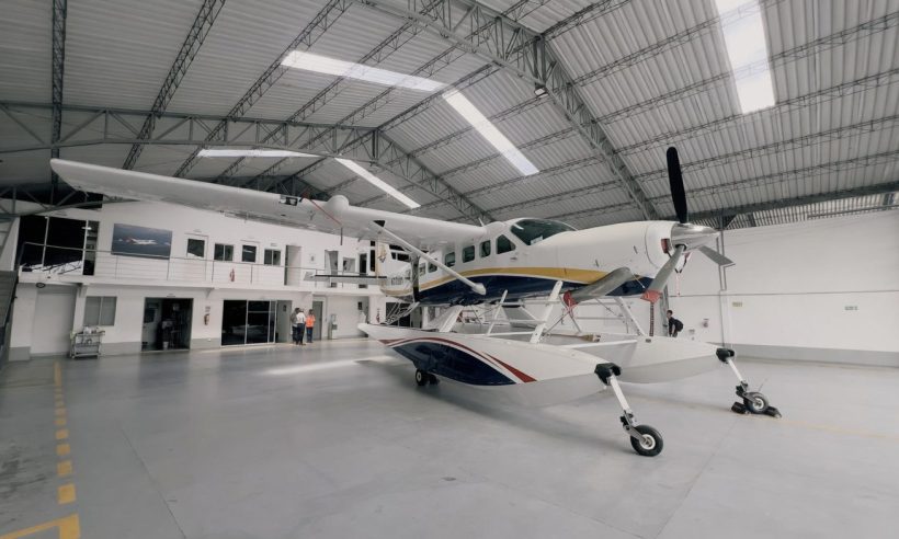A large white amphibious seaplane (Cessna Caravan-type aircraft with floats) is parked inside a spacious modern hangar. (Photo: Alma Air)