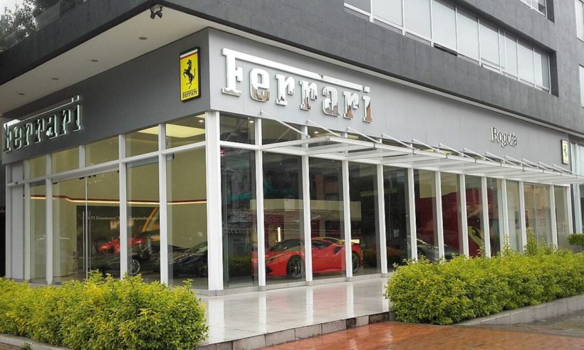 A modern glass Ferrari showroom with the iconic Ferrari logo and red sports cars visible inside, along with the landscaped entrance in Bogotá. (Photo: EEIM via Wikimedia Commons)