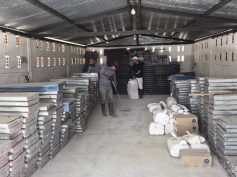 Metal trays of drill core are stacked in rows inside a storage shed while two workers sweep and tidy the floor.