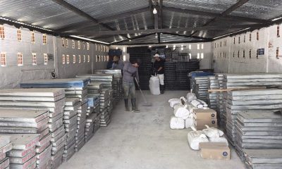 Metal trays of drill core are stacked in rows inside a storage shed while two workers sweep and tidy the floor.