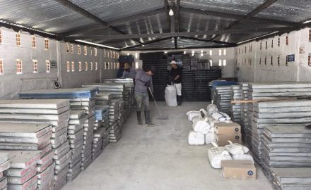Metal trays of drill core are stacked in rows inside a storage shed while two workers sweep and tidy the floor.