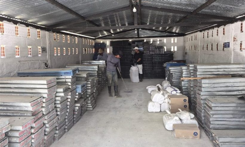 Metal trays of drill core are stacked in rows inside a storage shed while two workers sweep and tidy the floor.