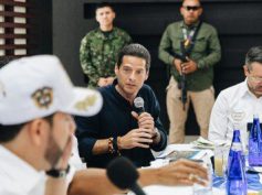Commerce, Industry and Tourism Minister Mauricio Gómez Amín speaks into a microphone as a military officer stands guard behind him.