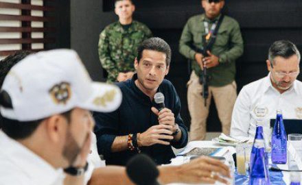 Commerce, Industry and Tourism Minister Mauricio Gómez Amín speaks into a microphone as a military officer stands guard behind him.