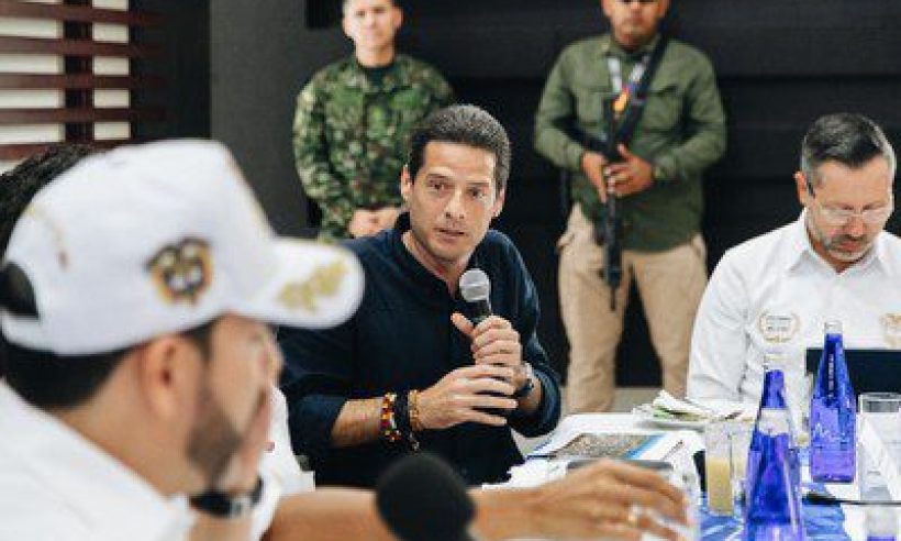 Commerce, Industry and Tourism Minister Mauricio Gómez Amín speaks into a microphone as a military officer stands guard behind him.