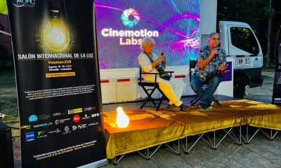 Two men holding microphones sit on an outdoor stage below a lit Cinemotion Labs screen and an ADFC festival banner.