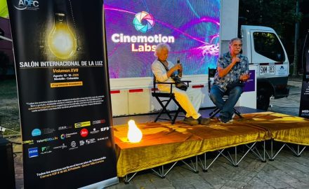 Two men holding microphones sit on an outdoor stage below a lit Cinemotion Labs screen and an ADFC festival banner.