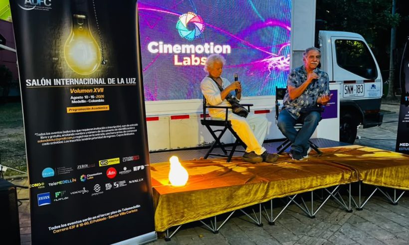 Two men holding microphones sit on an outdoor stage below a lit Cinemotion Labs screen and an ADFC festival banner.
