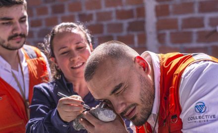 A man in a Collective Mining vest examines a rock sample with a hand lens while two others look on