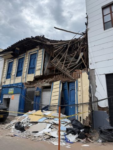 Two-story house with blue doors and a partly collapsed wood-and-bamboo roof, debris piled behind police tape