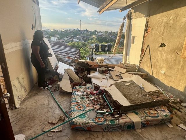 Woman stands in a bedroom where a collapsed wall buried a mattress in brick and tile, with a hillside town beyond