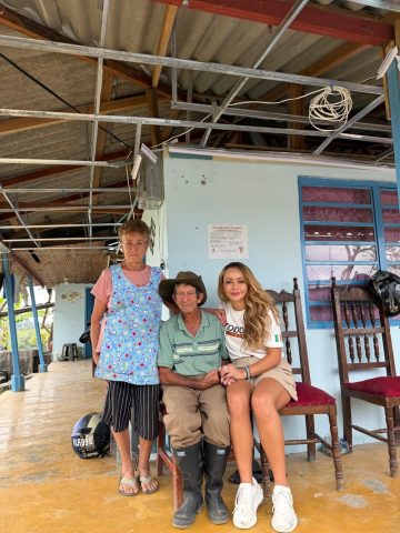Elderly couple sit with Mónica Gil Ryan on a porch with a damaged ceiling beside a posted danger-of-collapse notice