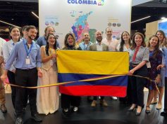 A group poses with a Colombian flag and a tricolor ribbon in front of a Colombia tourism map at a Paris trade fair stand.