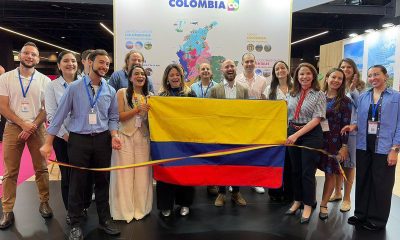 A group poses with a Colombian flag and a tricolor ribbon in front of a Colombia tourism map at a Paris trade fair stand.
