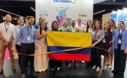 A group poses with a Colombian flag and a tricolor ribbon in front of a Colombia tourism map at a Paris trade fair stand.