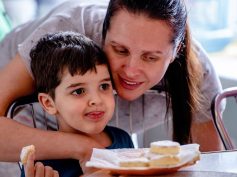 A mother embraces her young son at a cafe table set with pastries during breakfast.