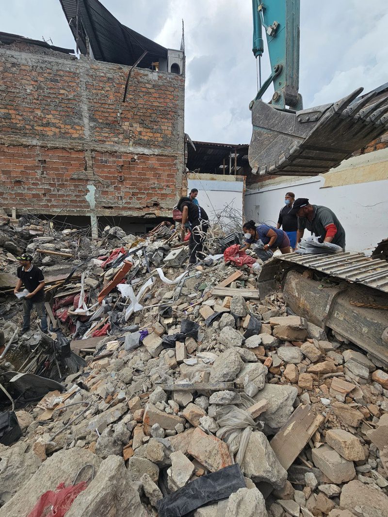 An excavator bucket looms over volunteers searching rubble beside a partially collapsed brick building.