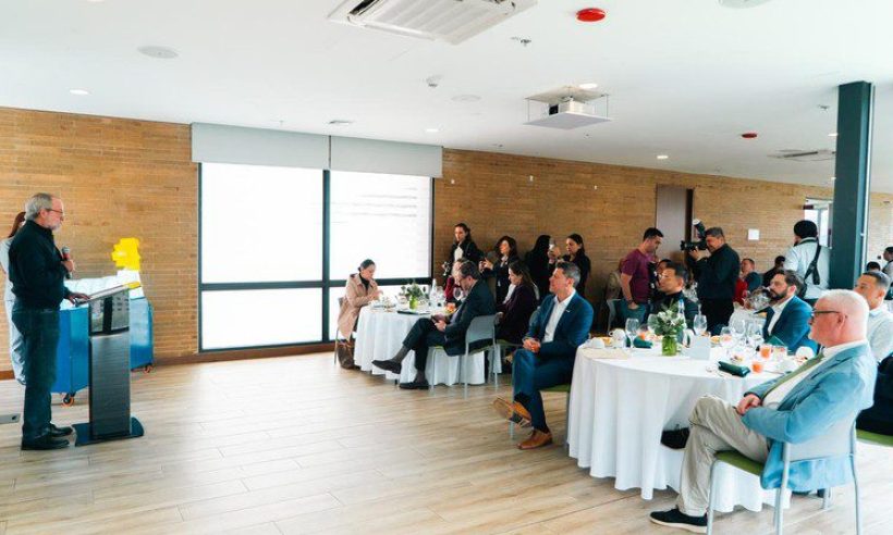 A speaker at a lectern addresses guests seated at round tables while photographers work at the back of the room.