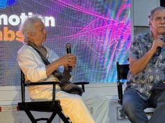 Two men are seated on stage in black director’s chairs during a panel at Cinemotion Labs (an audiovisual production and post-production event in Medellín, Colombia). The man on the left has white/gray curly hair, is dressed in a white shirt and white pants, and is holding a microphone. The man on the right has gray hair and a mustache, is wearing a blue patterned Hawaiian-style shirt, and is also holding a microphone.
