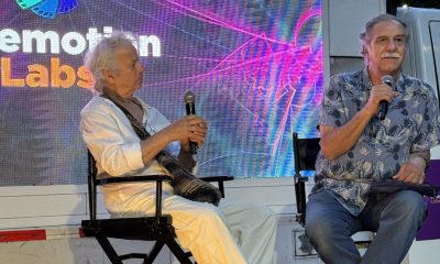 Two men are seated on stage in black director’s chairs during a panel at Cinemotion Labs (an audiovisual production and post-production event in Medellín, Colombia). The man on the left has white/gray curly hair, is dressed in a white shirt and white pants, and is holding a microphone. The man on the right has gray hair and a mustache, is wearing a blue patterned Hawaiian-style shirt, and is also holding a microphone.