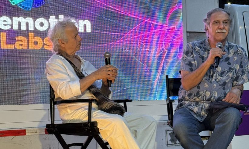 Two men are seated on stage in black director’s chairs during a panel at Cinemotion Labs (an audiovisual production and post-production event in Medellín, Colombia). The man on the left has white/gray curly hair, is dressed in a white shirt and white pants, and is holding a microphone. The man on the right has gray hair and a mustache, is wearing a blue patterned Hawaiian-style shirt, and is also holding a microphone.