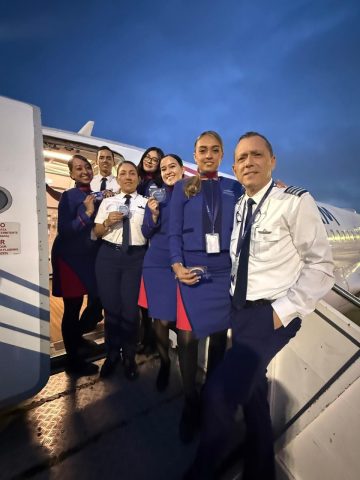 Seven LATAM Airlines Colombia pilots and flight attendants in uniform pose on the stairs at an aircraft door at dusk.
