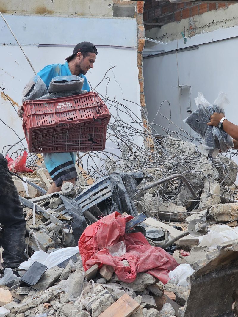 A man carries a plastic crate of salvaged goods through the wreckage of a collapsed building.