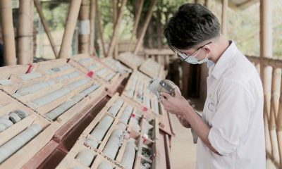 A masked worker holds a handheld instrument beside racks of drill core in wooden boxes under a bamboo-framed shed.