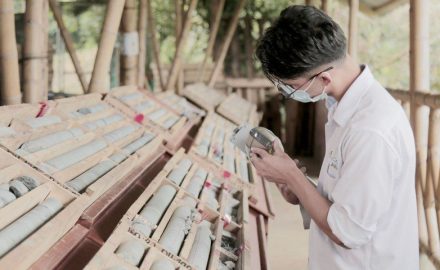 A masked worker holds a handheld instrument beside racks of drill core in wooden boxes under a bamboo-framed shed.