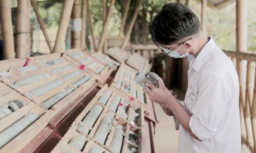 A masked worker holds a handheld instrument beside racks of drill core in wooden boxes under a bamboo-framed shed.