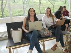 Two women sit on a bench working on laptops in a glass-walled hallway with trees visible outside.