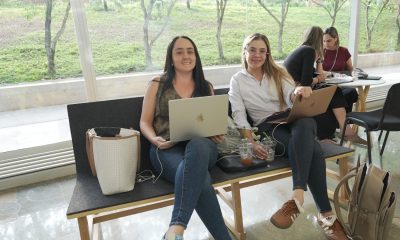 Two women sit on a bench working on laptops in a glass-walled hallway with trees visible outside.