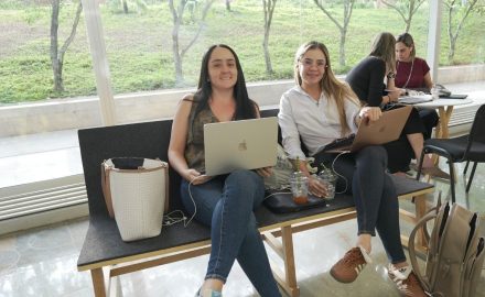 Two women sit on a bench working on laptops in a glass-walled hallway with trees visible outside.