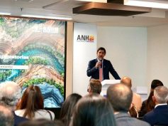 A speaker at a podium in front of an ANH banner addresses an audience beside a screen showing an exploration presentation