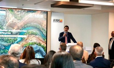 A speaker at a podium in front of an ANH banner addresses an audience beside a screen showing an exploration presentation