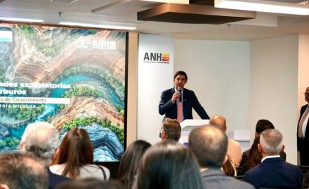 A speaker at a podium in front of an ANH banner addresses an audience beside a screen showing an exploration presentation