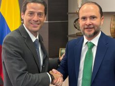Two men in suits shake hands in an office in front of a Colombian flag.