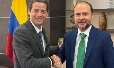 Two men in suits shake hands in an office in front of a Colombian flag.