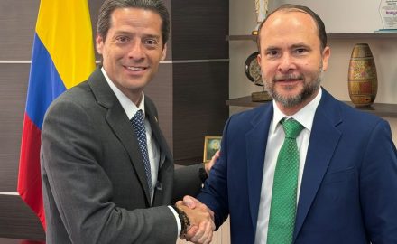 Two men in suits shake hands in an office in front of a Colombian flag.