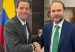 Two men in suits shake hands in an office in front of a Colombian flag.