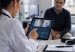 A physician reviews remote patient monitoring data on a tablet during a telemedicine consultation with a patient present in the office
