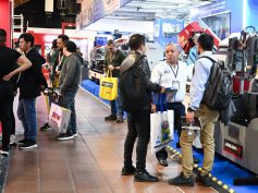 Visitors walk through a trade fair aisle lined with metalworking machinery, including industrial band saws