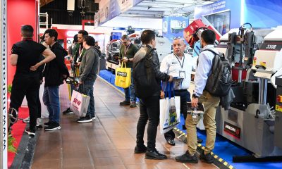 Visitors walk through a trade fair aisle lined with metalworking machinery, including industrial band saws