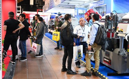 Visitors walk through a trade fair aisle lined with metalworking machinery, including industrial band saws