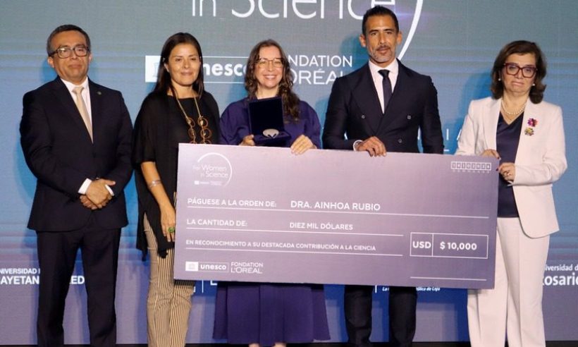 Five people stand on a stage before a For Women in Science backdrop, one holding an award medal in a case.