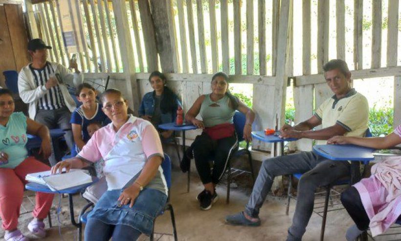 Adults seated at blue desk-chairs talk inside the wooden classroom