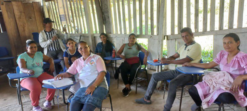 Adults seated at blue desk-chairs talk inside the wooden classroom